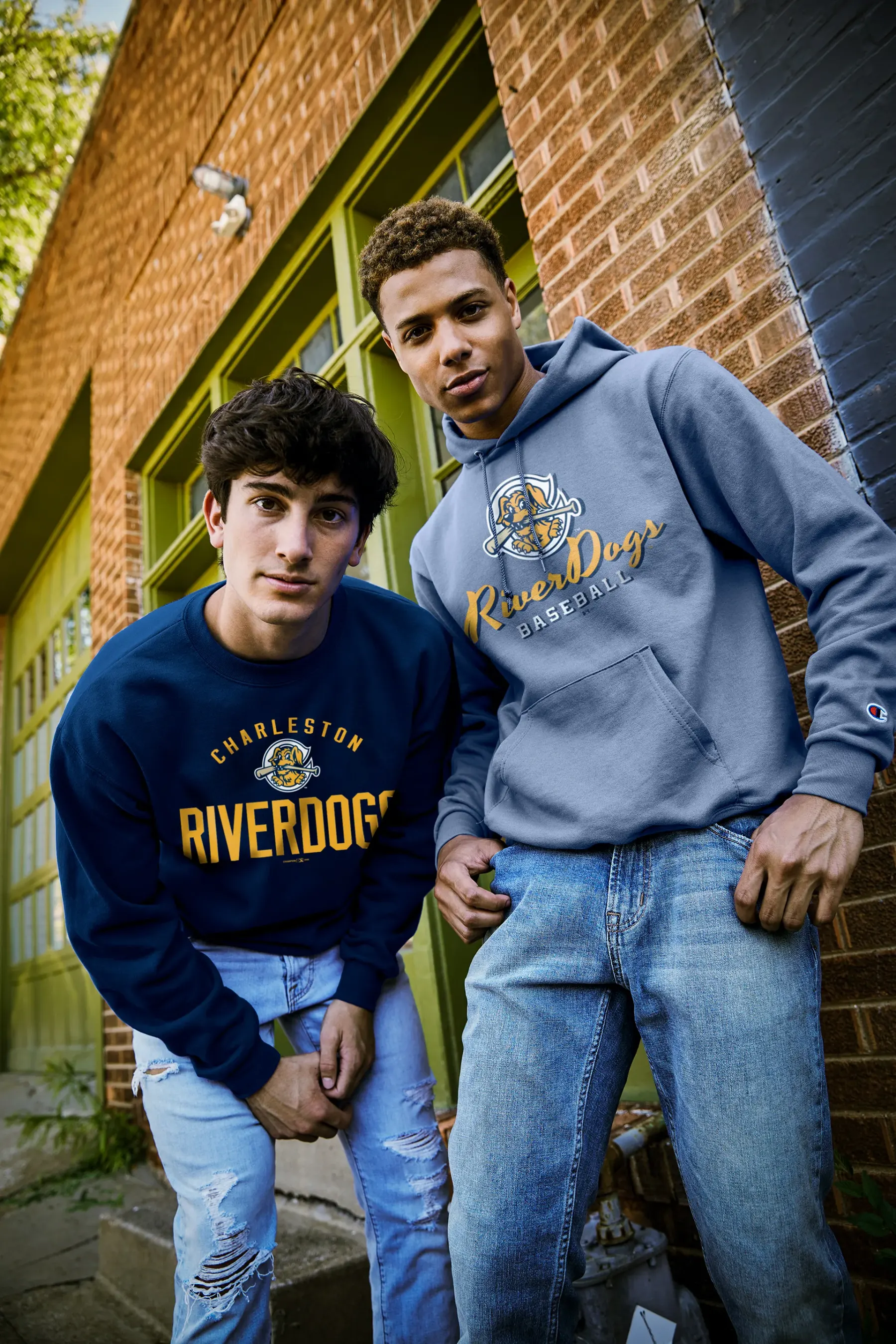 Two men against a brick and green-painted wall in navy and pale blue collegiate sweatshirts and jeans, one carrying a Charleston RiverDogs crest and the other a RiverDogs script across the chest.