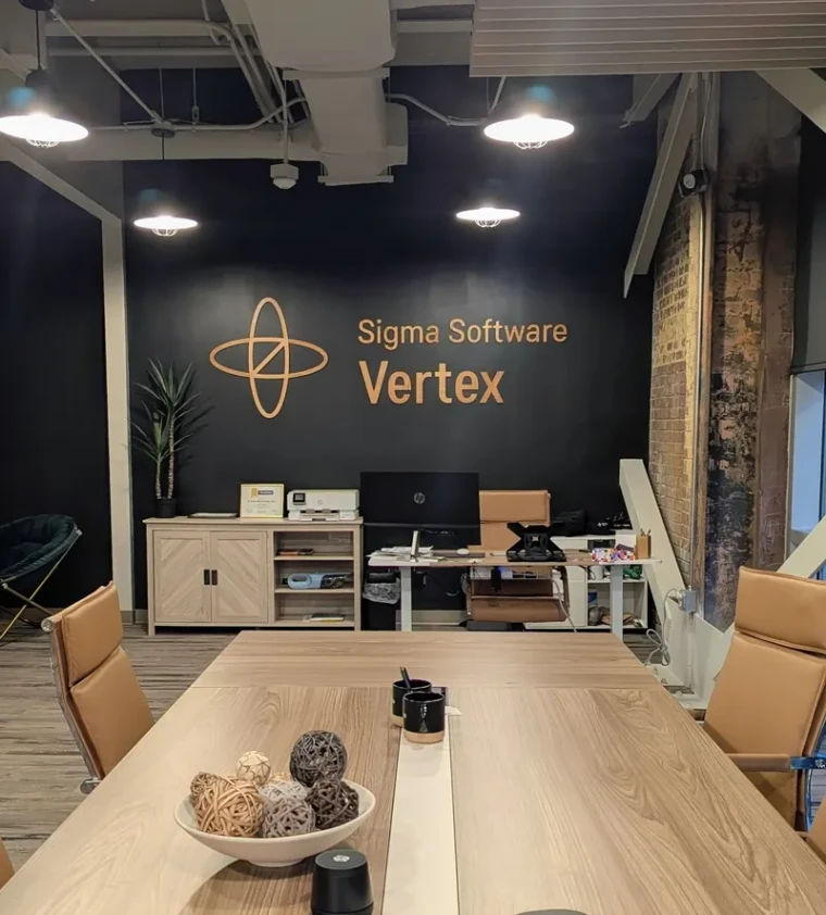 A meeting room with exposed ceiling services and a long pale table, with the Sigma Software Vertex mark and lettering mounted on the dark end wall.