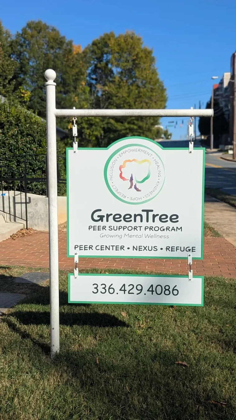 A white post-mounted sign in a grass verge reading GreenTree Peer Support Program, with the rainbow tree mark, the strapline Growing Mental Wellness, and a phone number.