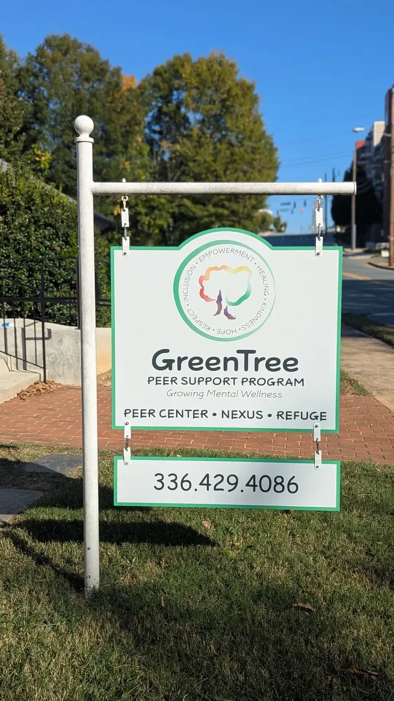 A white post-mounted sign in a grass verge reading GreenTree Peer Support Program, with the rainbow tree mark, the strapline Growing Mental Wellness, and a phone number.