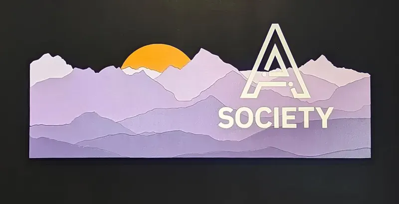 A layered wooden sign on black: a purple mountain range in stepped layers with a sun rising behind it, and the A Society mark cut into the ridgeline.