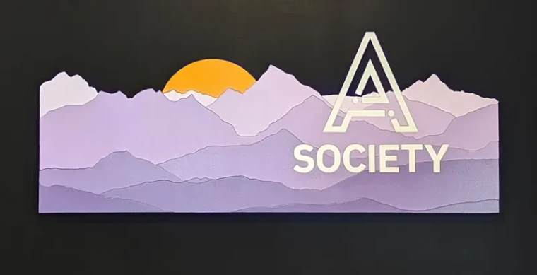 A layered wooden sign on black: a purple mountain range in stepped layers with a sun rising behind it, and the A Society mark cut into the ridgeline.