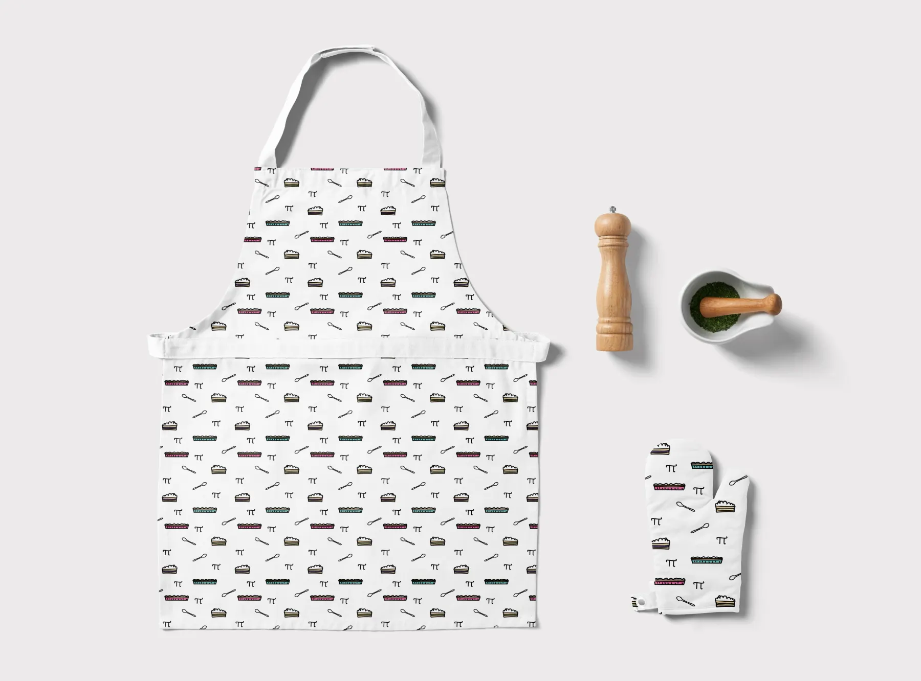The pattern printed on a white apron and a matching oven glove laid flat on pale grey, with a wooden pepper mill and a mortar of chopped herbs alongside.