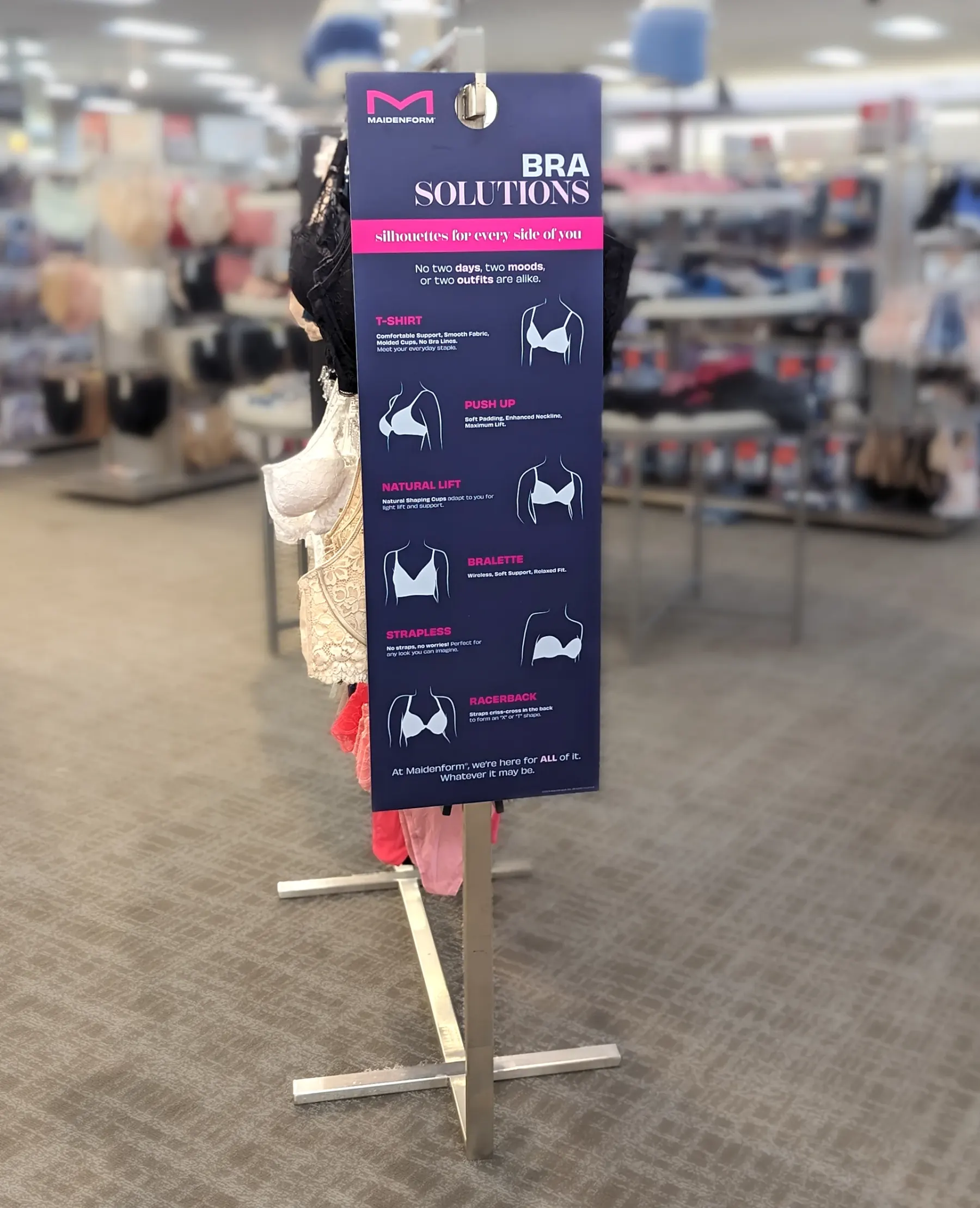 The Bra Solutions panel printed and mounted on a steel floor stand in a department store, bras on a rail behind it and the shop floor out of focus beyond.